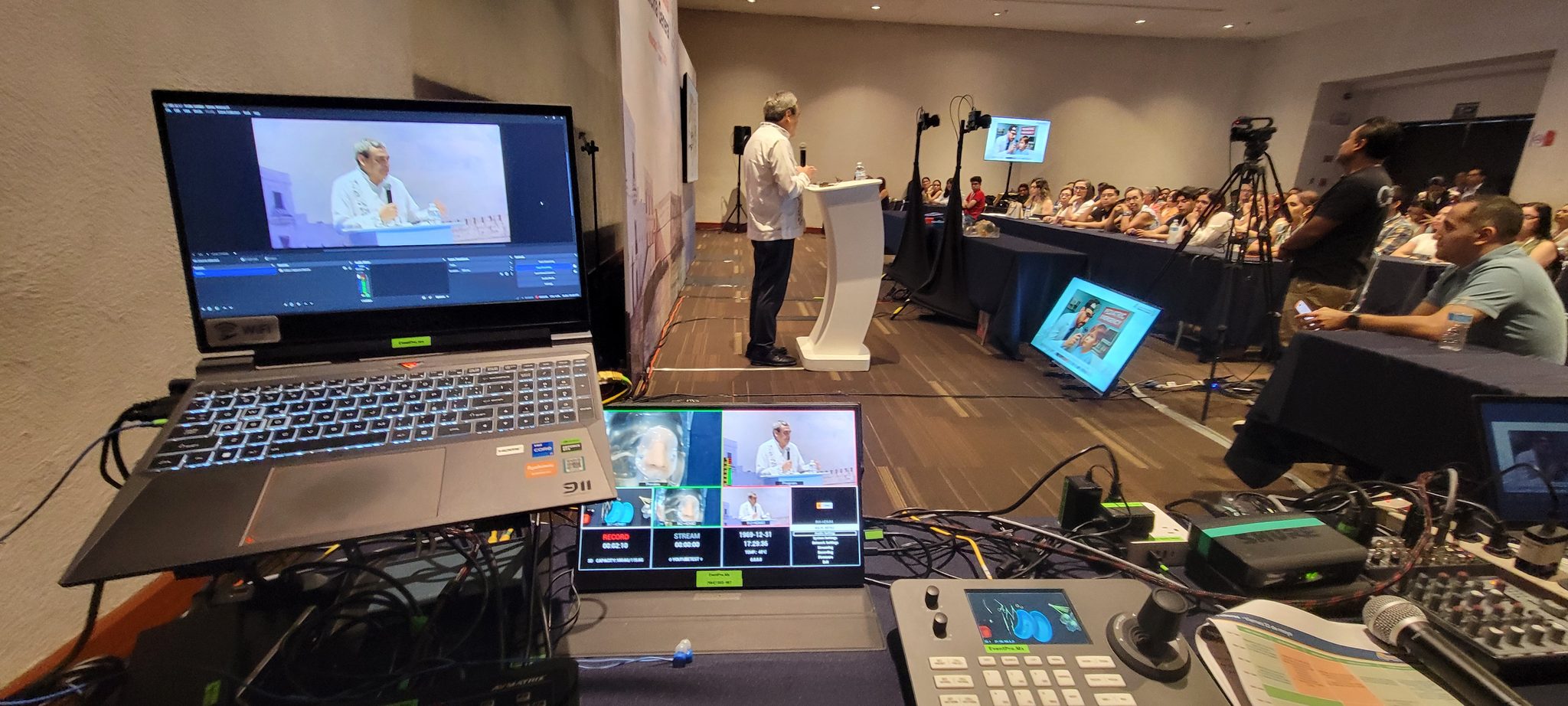Estación de monitoreo y streaming en primer plano con ponente y audiencia de conferencia al fondo
