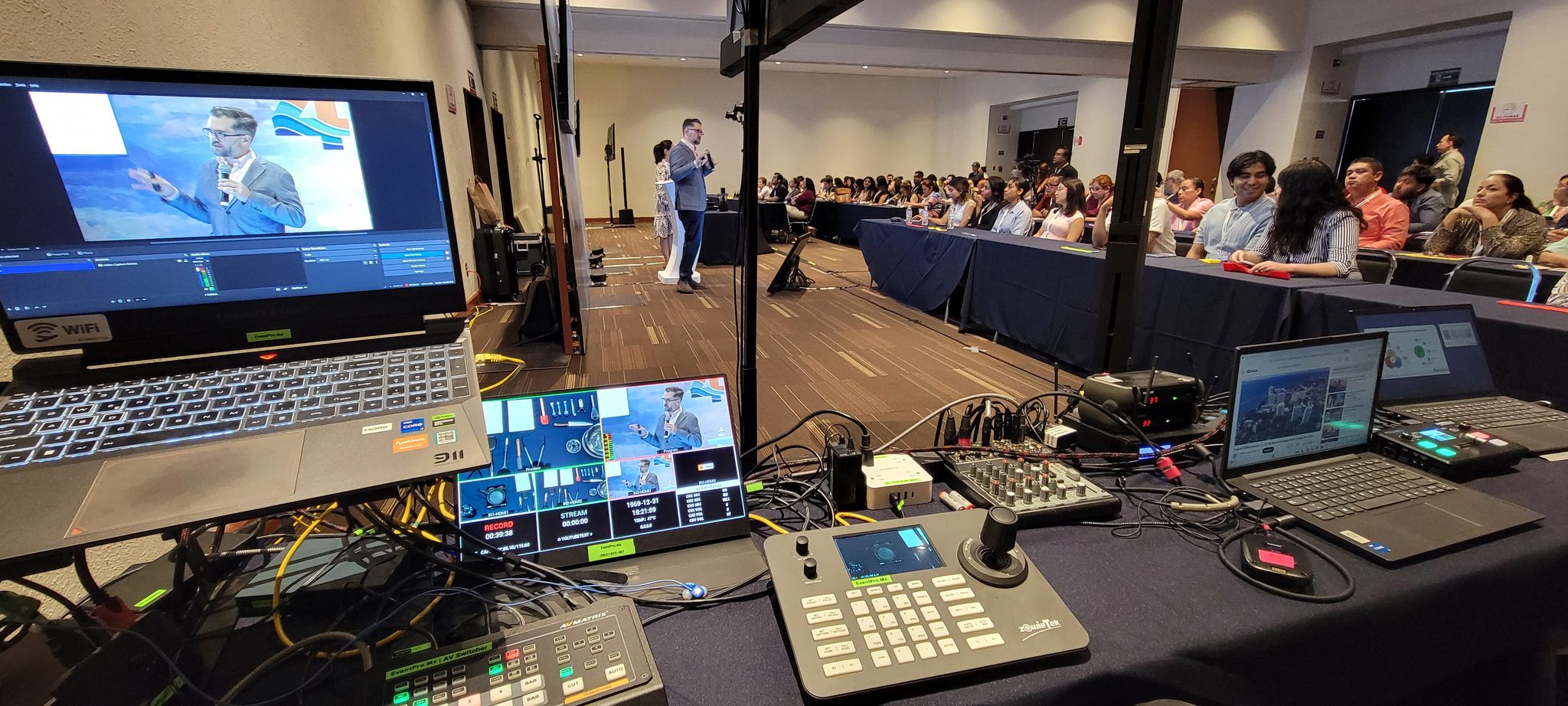 Mesa de producción multicámara con control PTZ, laptops y monitores durante una conferencia con audiencia
