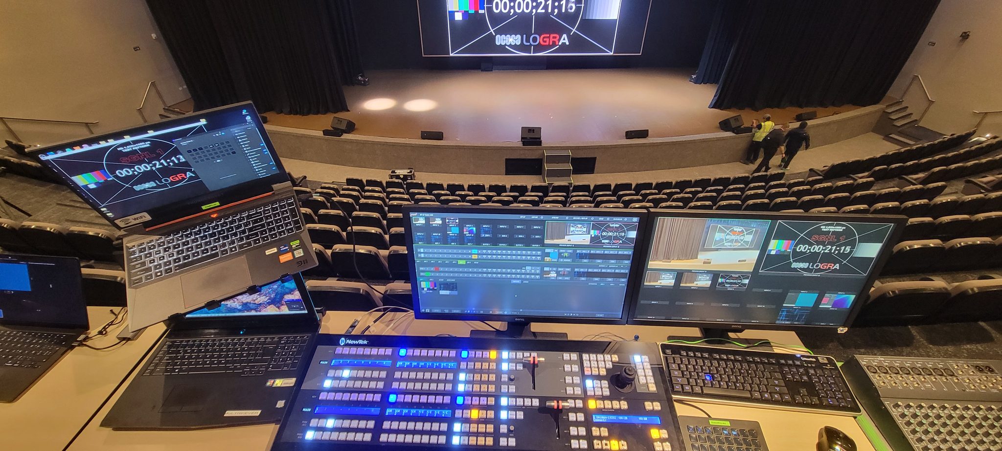 Mesa de control con switcher de video iluminado y monitores de producción frente a un teatro con pantalla LED
